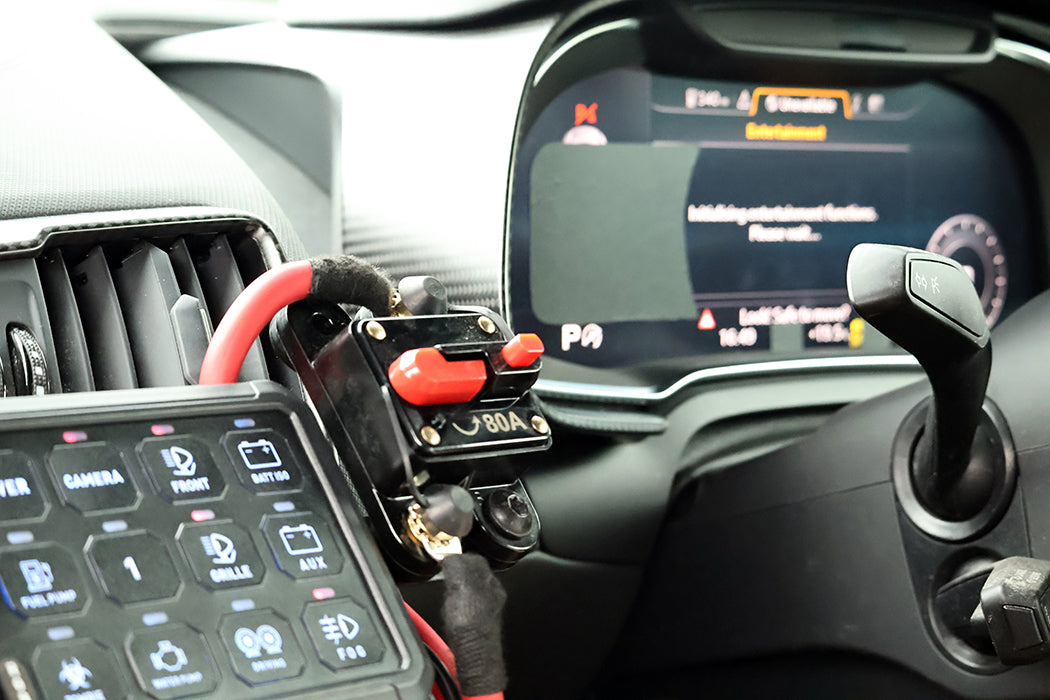 A close-up of a car's dashboard and center console showing a custom black electrical switch with red buttons and red/black wiring connected near the infotainment screen and gear shift lever.